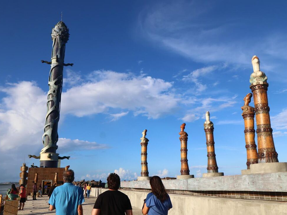 Grupo de pessoas observando estátuas altas e ornamentadas contra um fundo de céu azul brilhante, capturando a interseção da curiosidade humana e da expressão artística.