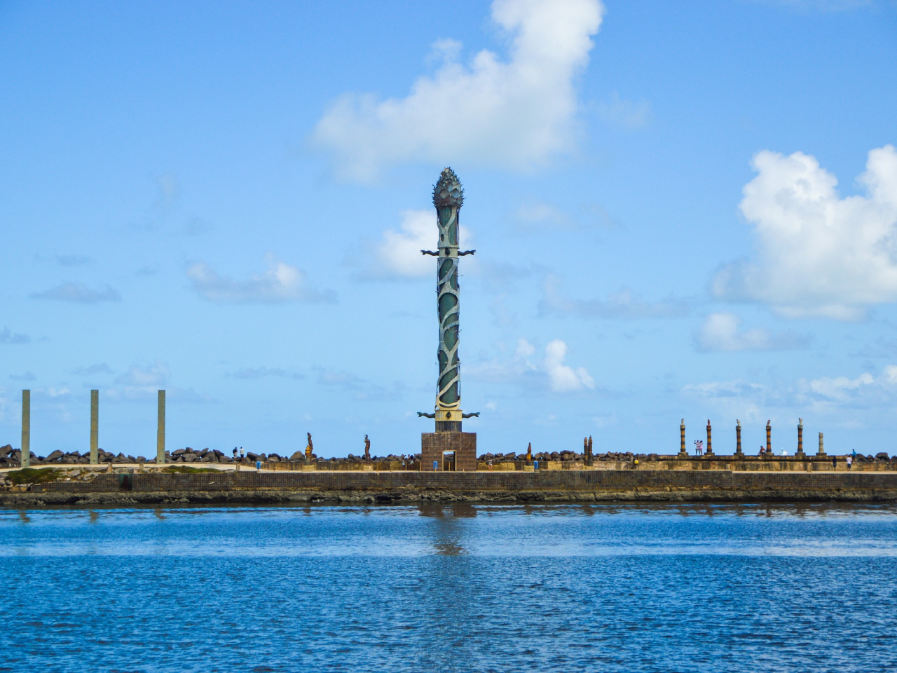 Monumento artístico alto situado em meio a um corpo de água azul sereno, sob o céu claro com nuvens dispersas, criando uma cena pitoresca pela harmoniosa combinação de arte humana e beleza natural.